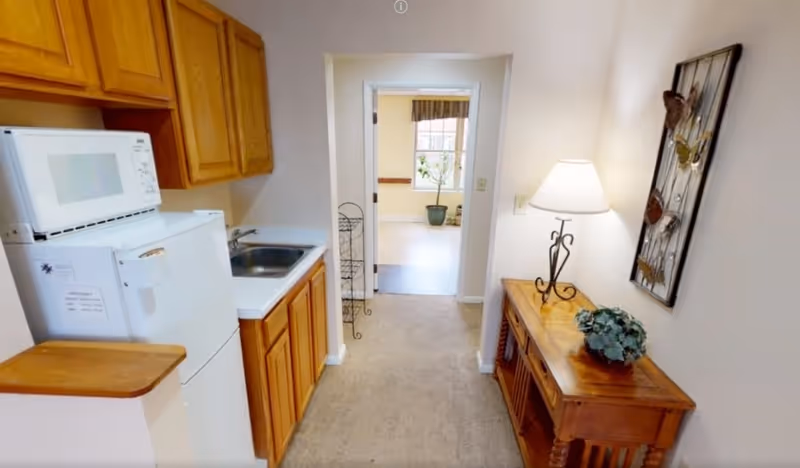 Small kitchenette and hallway with a refrigerator, microwave, sink and wooden cabinets opposite a console table with a lamp leading to another room.