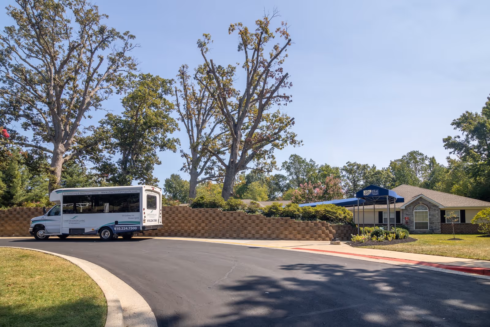 Exterior view of Charter Senior Living of Annapolis showing a driveway with a white shuttle bus parked on the left side. The building is partially visible on the right with a blue awning over the entrance. There are trees and landscaping around the area under a clear blue sky.
