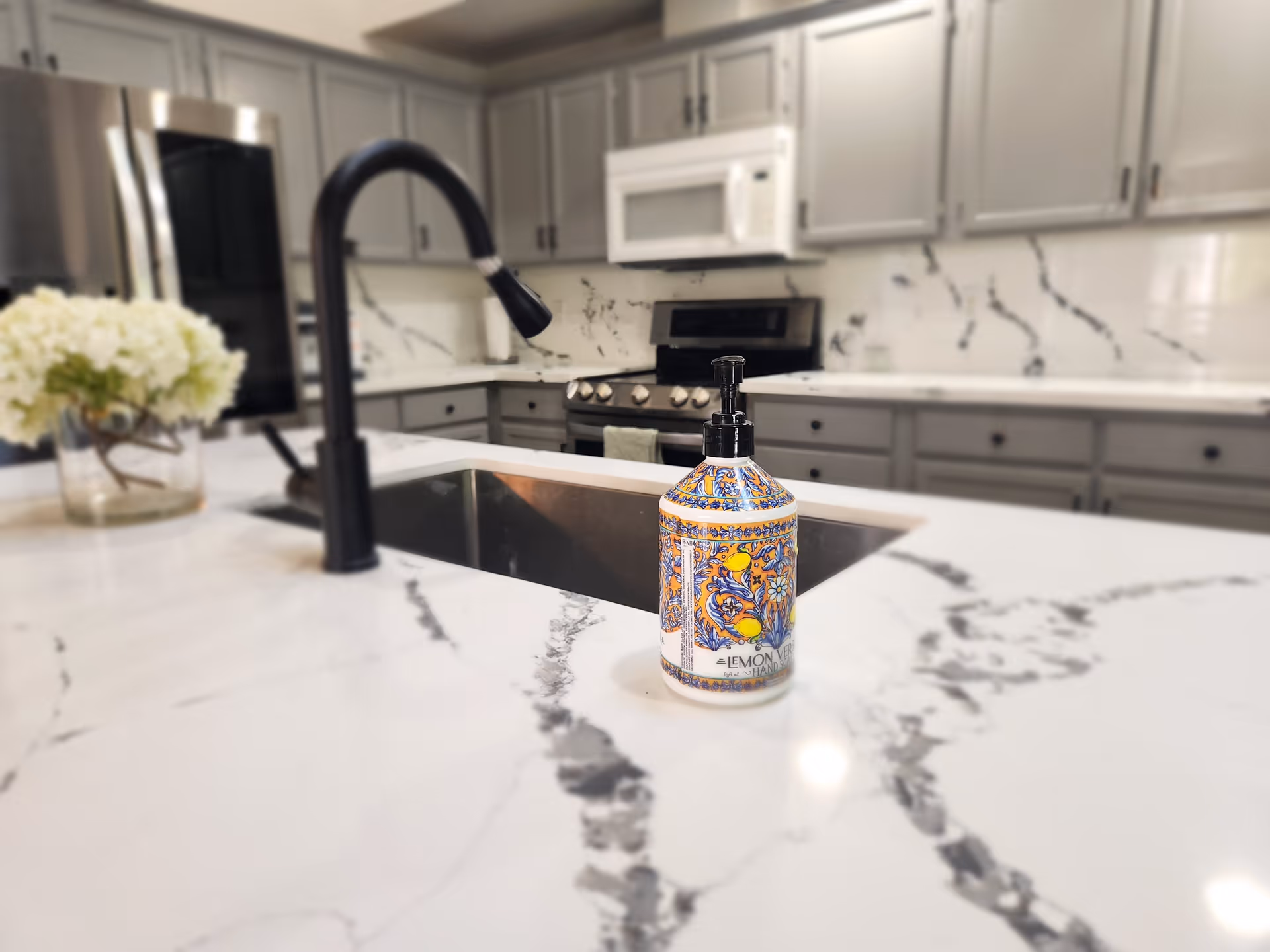 Modern kitchen island with a black faucet, patterned soap dispenser on a white marble countertop and cabinets and appliances in the background.