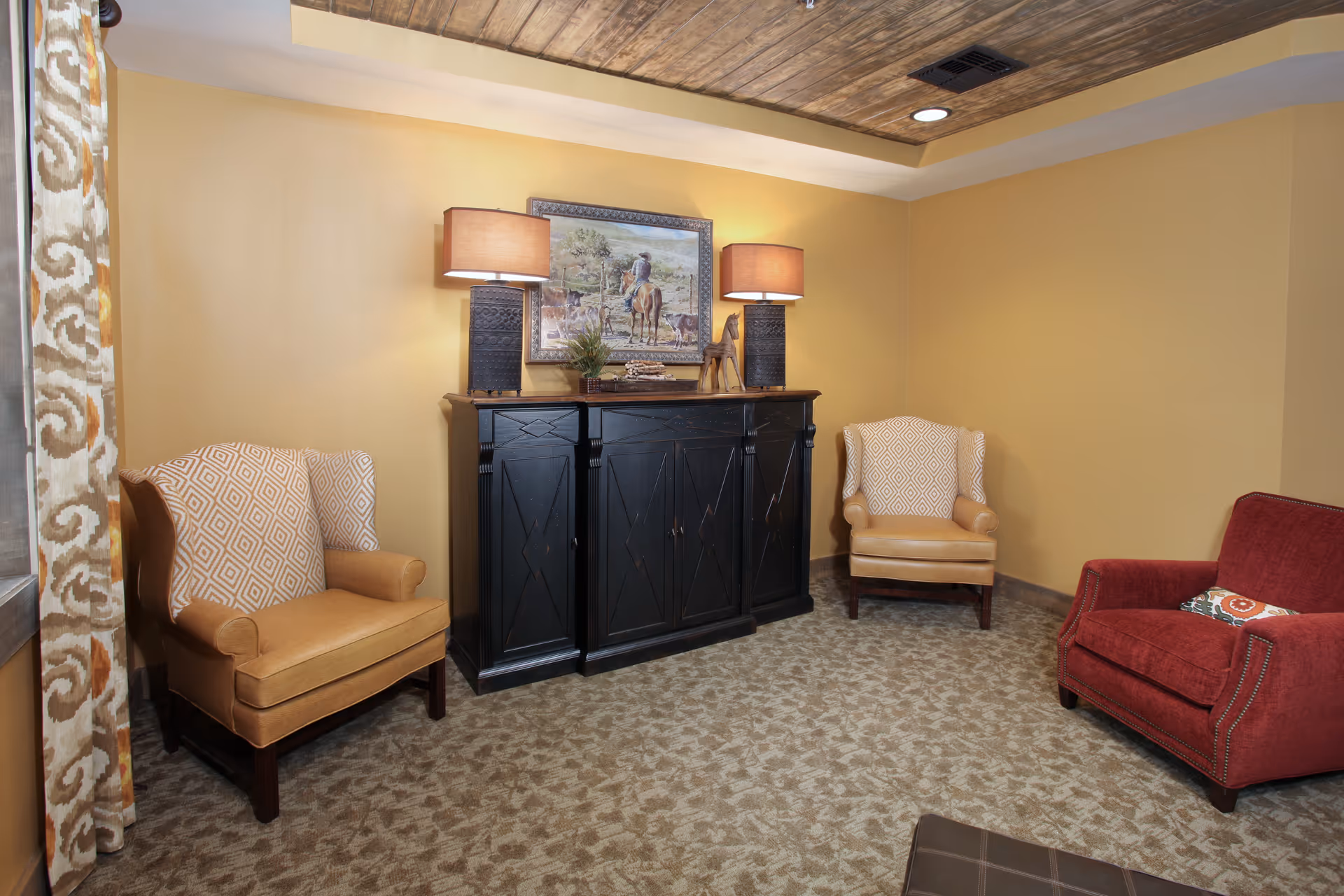 A cozy sitting area with two beige armchairs featuring patterned cushions, a red armchair with a decorative pillow, and a black wooden cabinet with two table lamps and a framed painting of a person riding a horse on the wall above it. The room has yellow walls, a patterned carpet, and a wooden ceiling panel with recessed lighting.