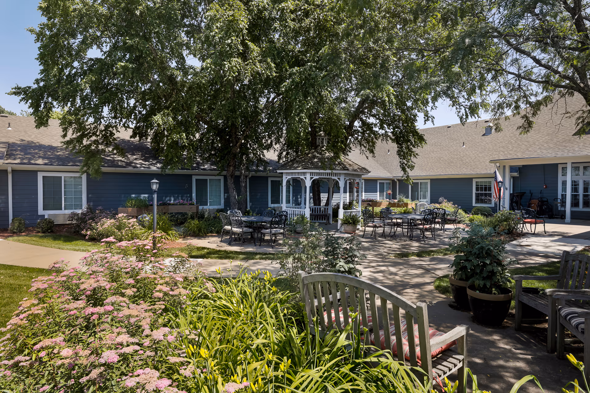 Outdoor courtyard area of a senior living facility with a large tree providing shade over a white gazebo surrounded by metal tables and chairs. There are flower beds with pink flowers and green plants, wooden benches with cushions, and a blue building in the background under a clear sky.