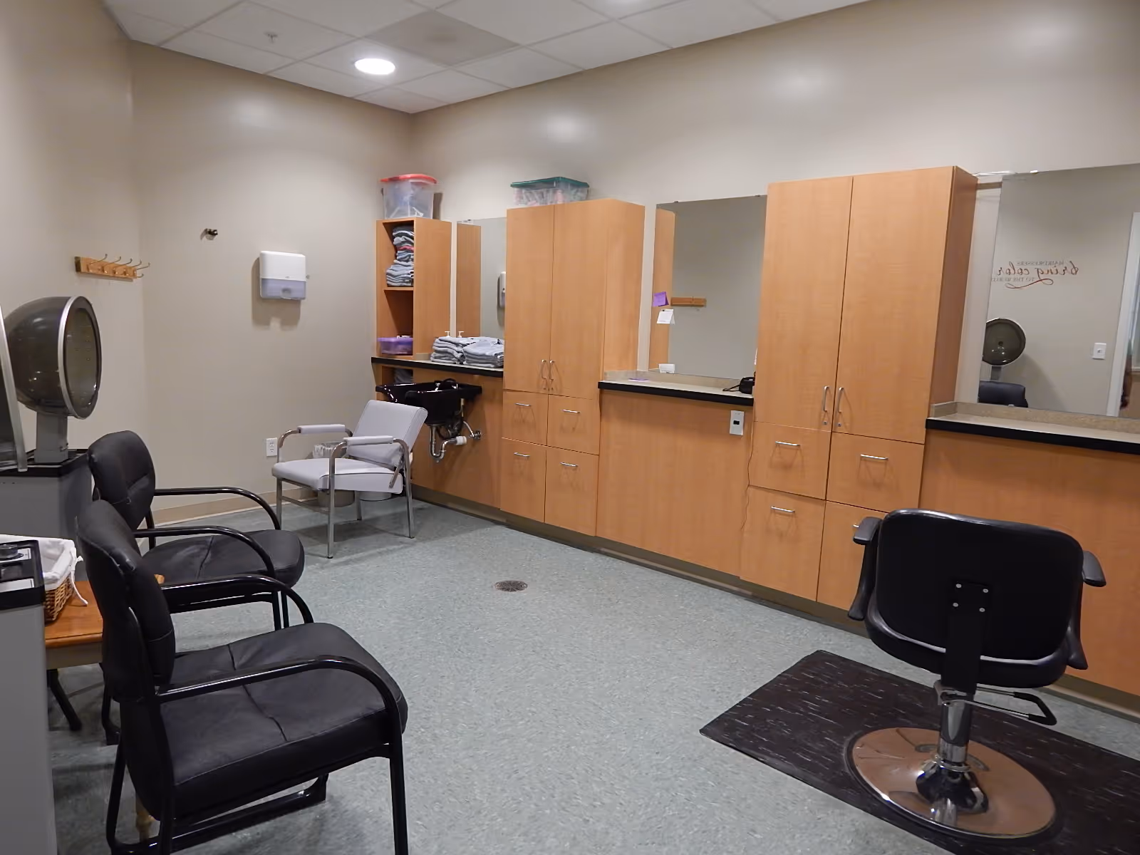 A hair-salon style room with barber chairs, a hooded dryer, sinks, towels, and wooden cabinets along the wall.