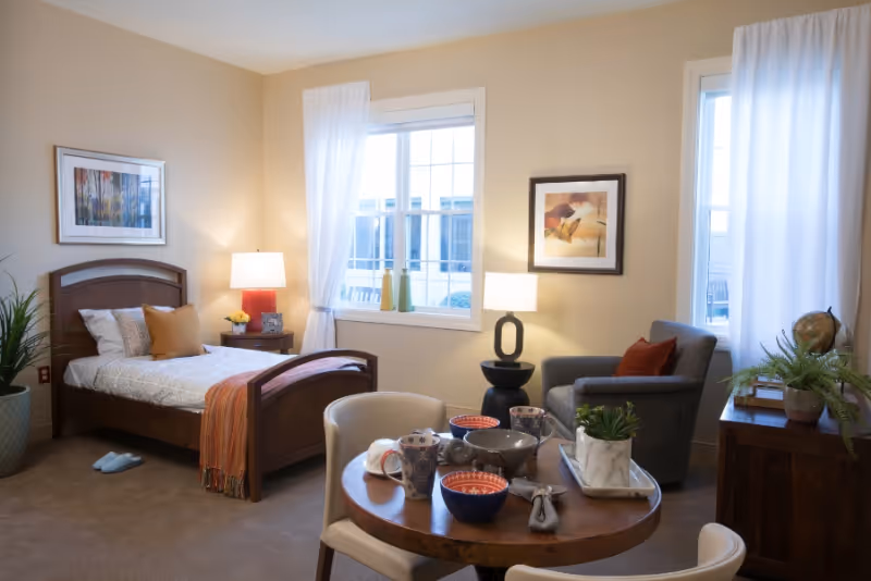 A bright studio room with a single bed, bedside table and lamp, round dining table set with dishes, an armchair, and windows with white curtains.