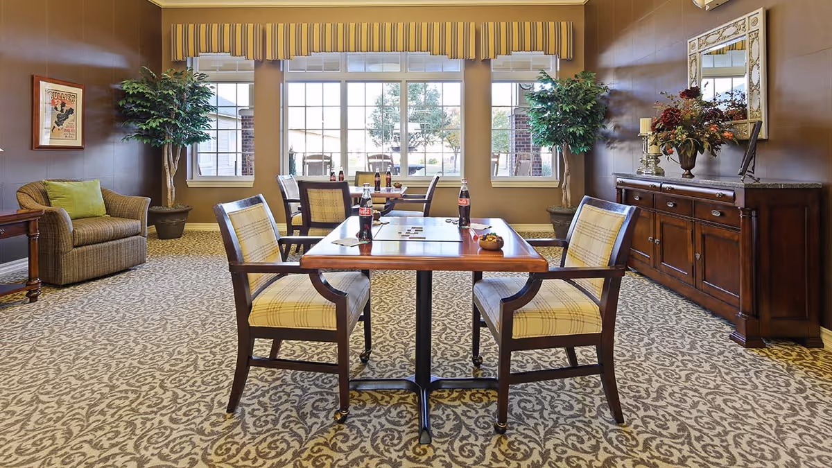 A well-lit common area with patterned carpet, featuring a wooden table with four cushioned chairs around it. On the table are several bottles of Coca-Cola and some small bowls. The room has large windows with striped valances, two large potted plants, a comfortable armchair with a green pillow, a wooden sideboard with decorative items including a vase with flowers, candles, and a large mirror above it. The walls are painted in warm tones.