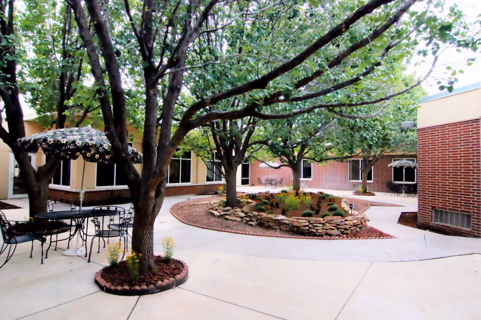 Outdoor courtyard area with several trees, a circular stone planter with flowers, and metal tables and chairs with floral-patterned umbrellas. The courtyard is surrounded by brick and beige building walls with windows.