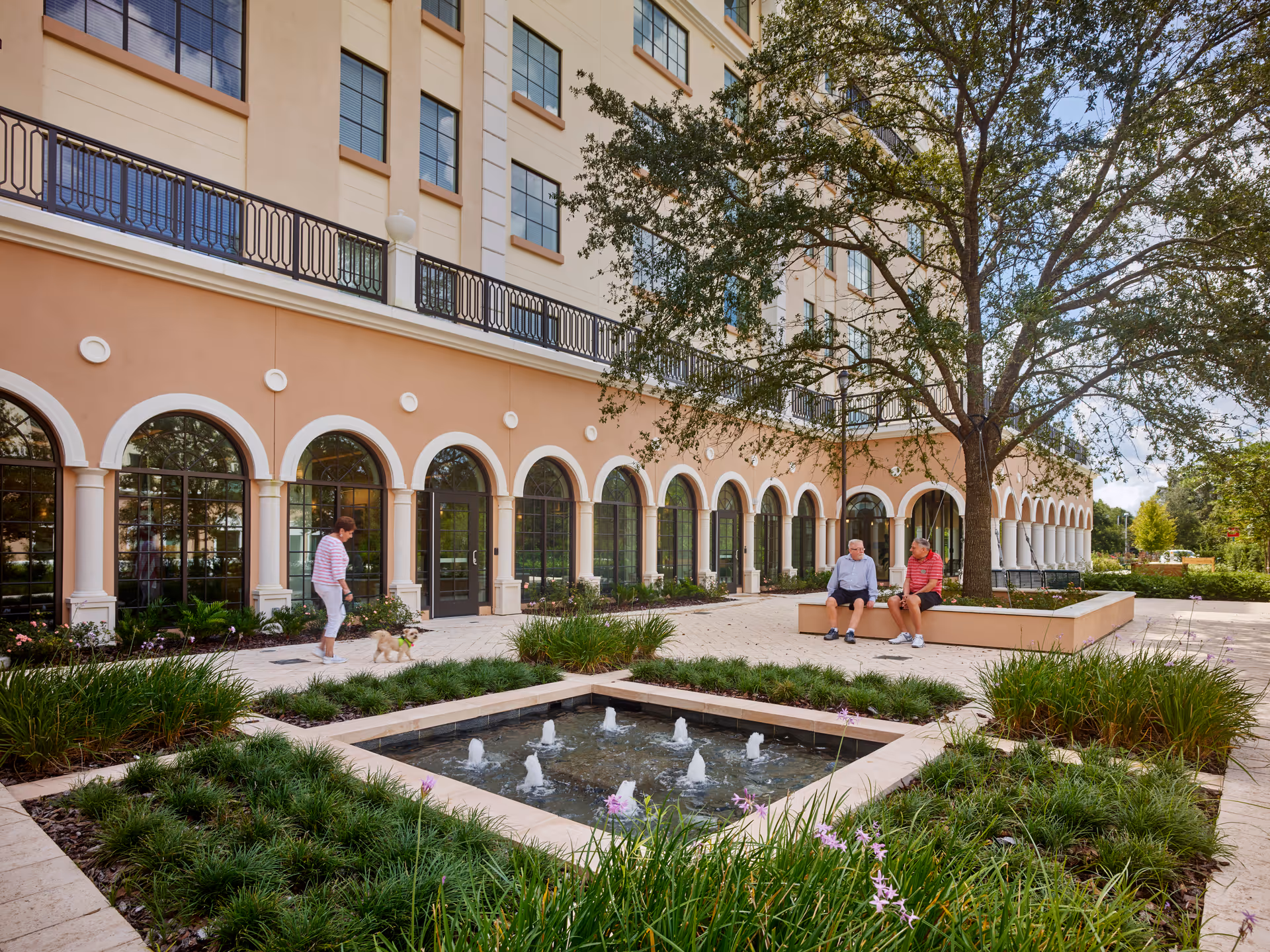 Outdoor courtyard area of a senior living facility with a square water fountain surrounded by greenery and flowers. Two elderly men are sitting on a bench under a large tree, and a woman is walking a small dog along the pathway. The building in the background has large arched windows and a beige facade.