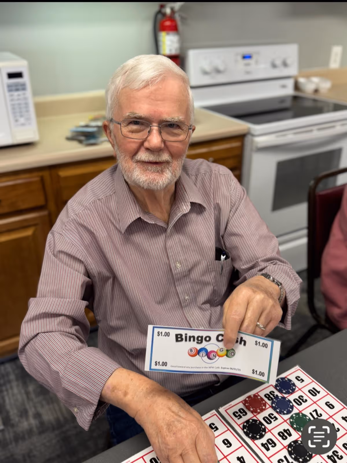An elderly man with white hair and glasses sitting at a table in a kitchen area, holding a Bingo cash prize ticket and smiling. A Bingo card with chips is on the table in front of him. Behind him are kitchen cabinets, a microwave, a stove, and a fire extinguisher.