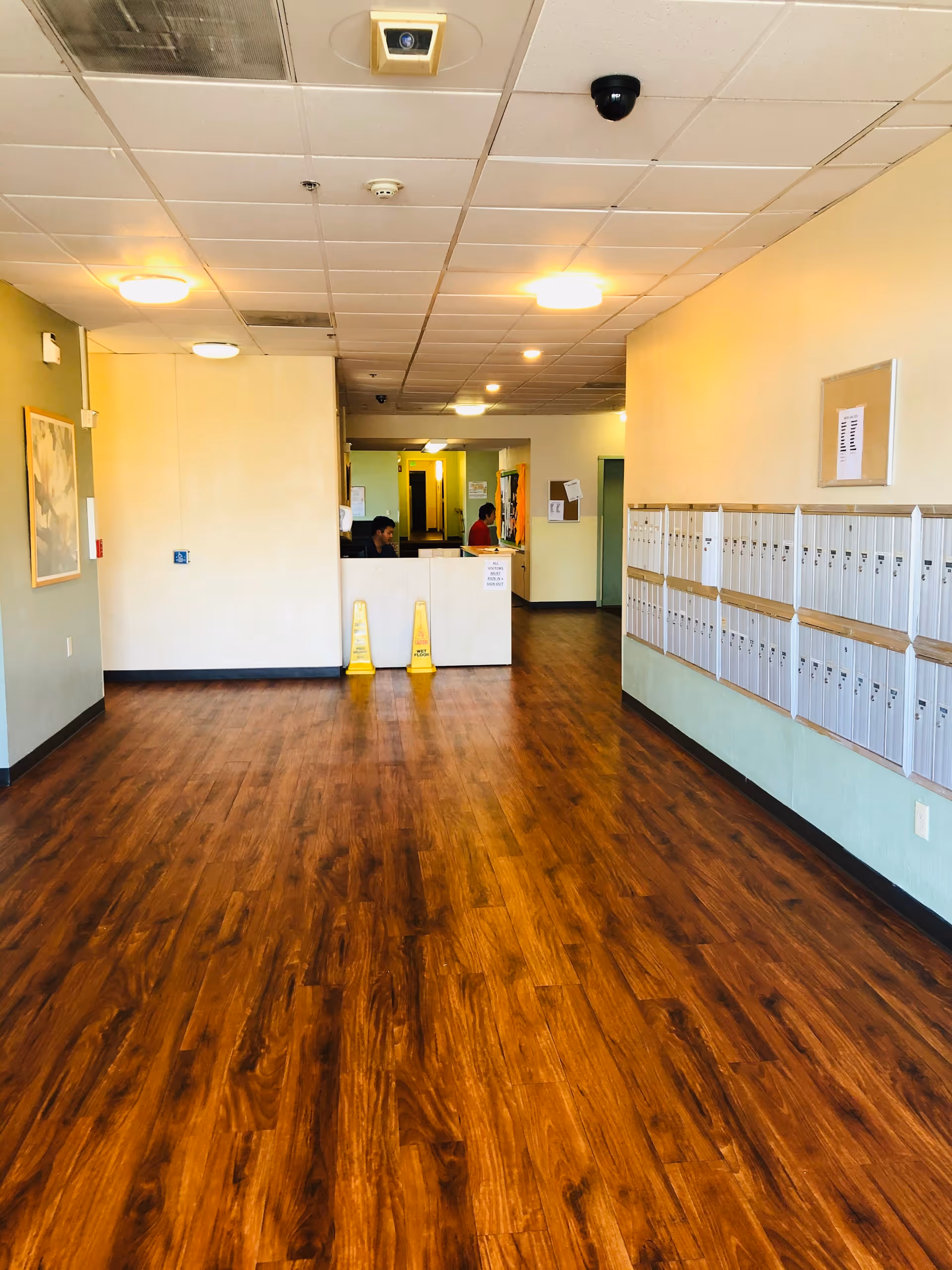 Interior hallway of a senior living facility with wood flooring, a row of mailboxes on the right wall, a reception desk with two staff members, and yellow caution wet floor signs in front of the desk.
