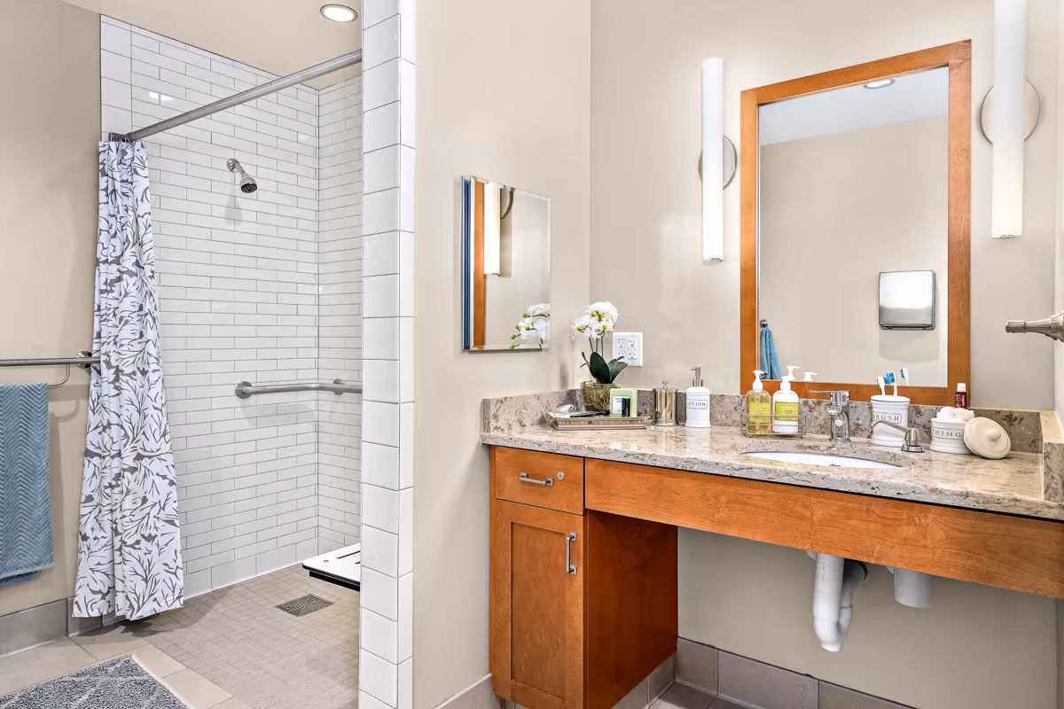 Accessible bathroom with a tiled walk-in shower, shower curtain, grab bars, and a vanity with sink, mirror, and toiletries.