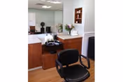 Interior view of a salon area within a senior living facility featuring a black salon chair in front of a wooden cabinet with a sink, a large mirror, and shelves holding towels and flowers.