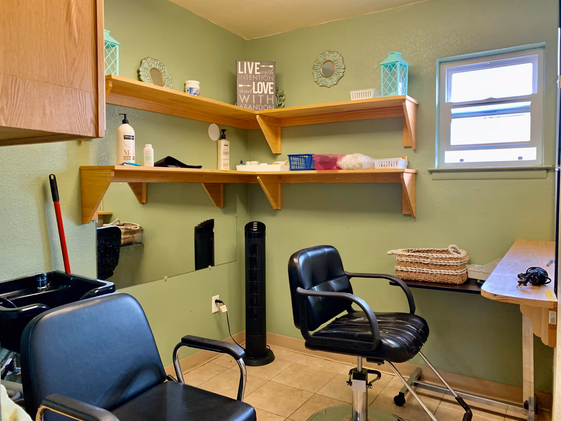 A small salon or grooming room with two black salon chairs, a black sink, wooden shelves holding various items including bottles, baskets, and decorative pieces, a small window letting in natural light, and a standing fan against a green wall.