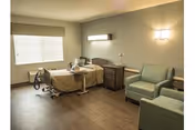 A well-lit nursing facility room with a hospital bed, bedside table, two green armchairs, and a large window with blinds. The room has wooden flooring and neutral-colored walls with modern light fixtures.