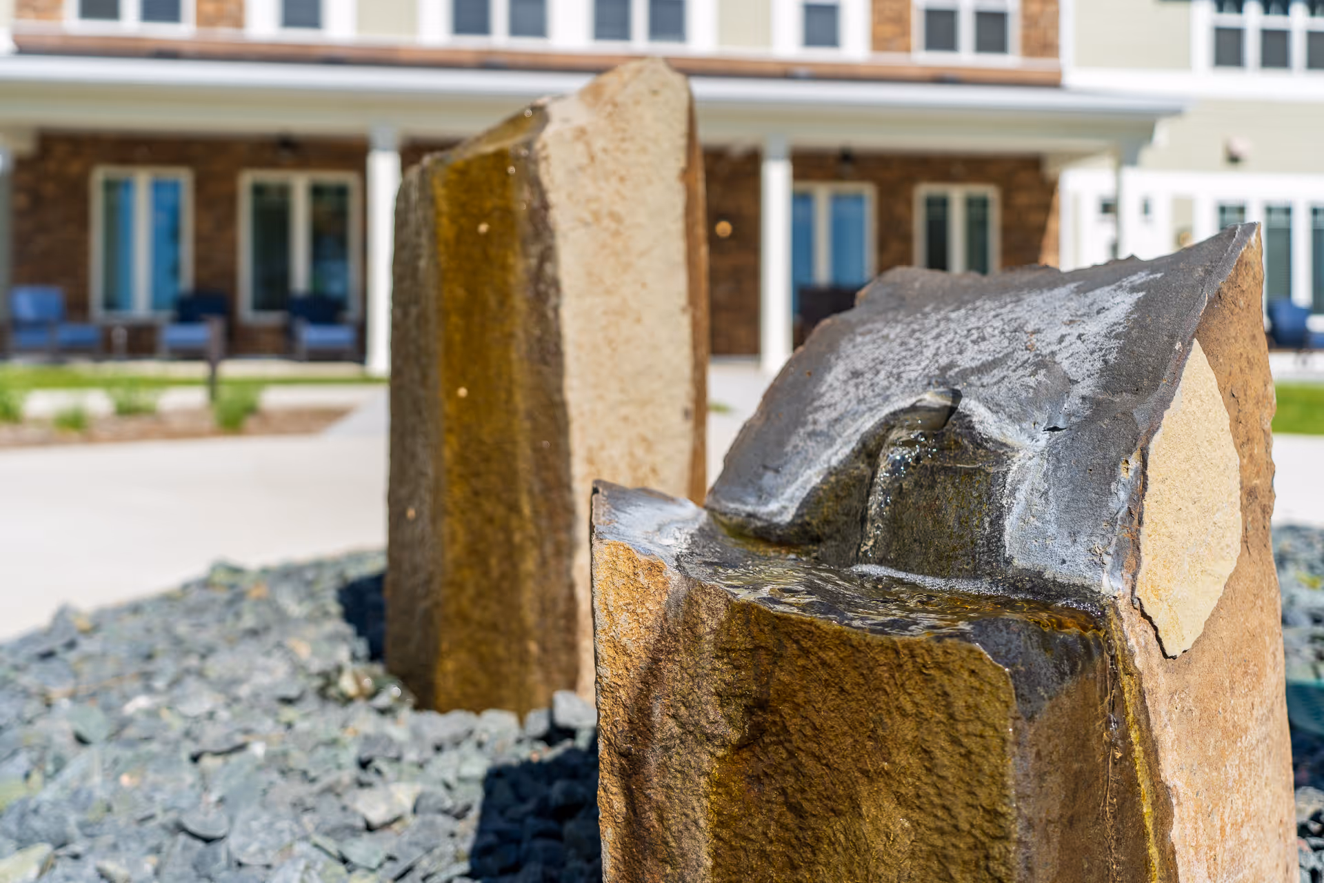 Close-up of a stone water fountain in a courtyard with the senior living building and porch seating blurred in the background.