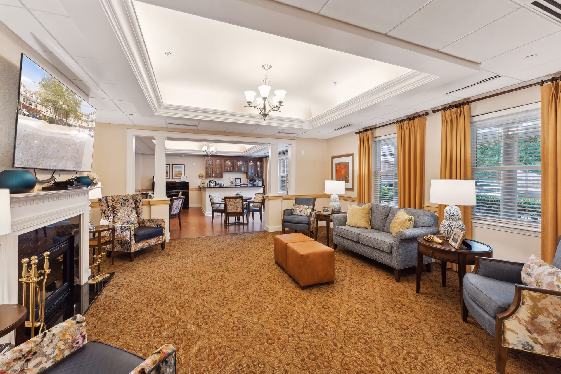 A cozy living room area in a senior living facility featuring a patterned carpet, a gray sofa with yellow pillows, two armchairs with floral upholstery, two side tables with lamps and framed photos, large windows with mustard yellow curtains, a fireplace with decorative tools, and a wall-mounted TV displaying an outdoor scene. In the background, there is a dining area with a table and chairs and a kitchen with wooden cabinets and a counter.