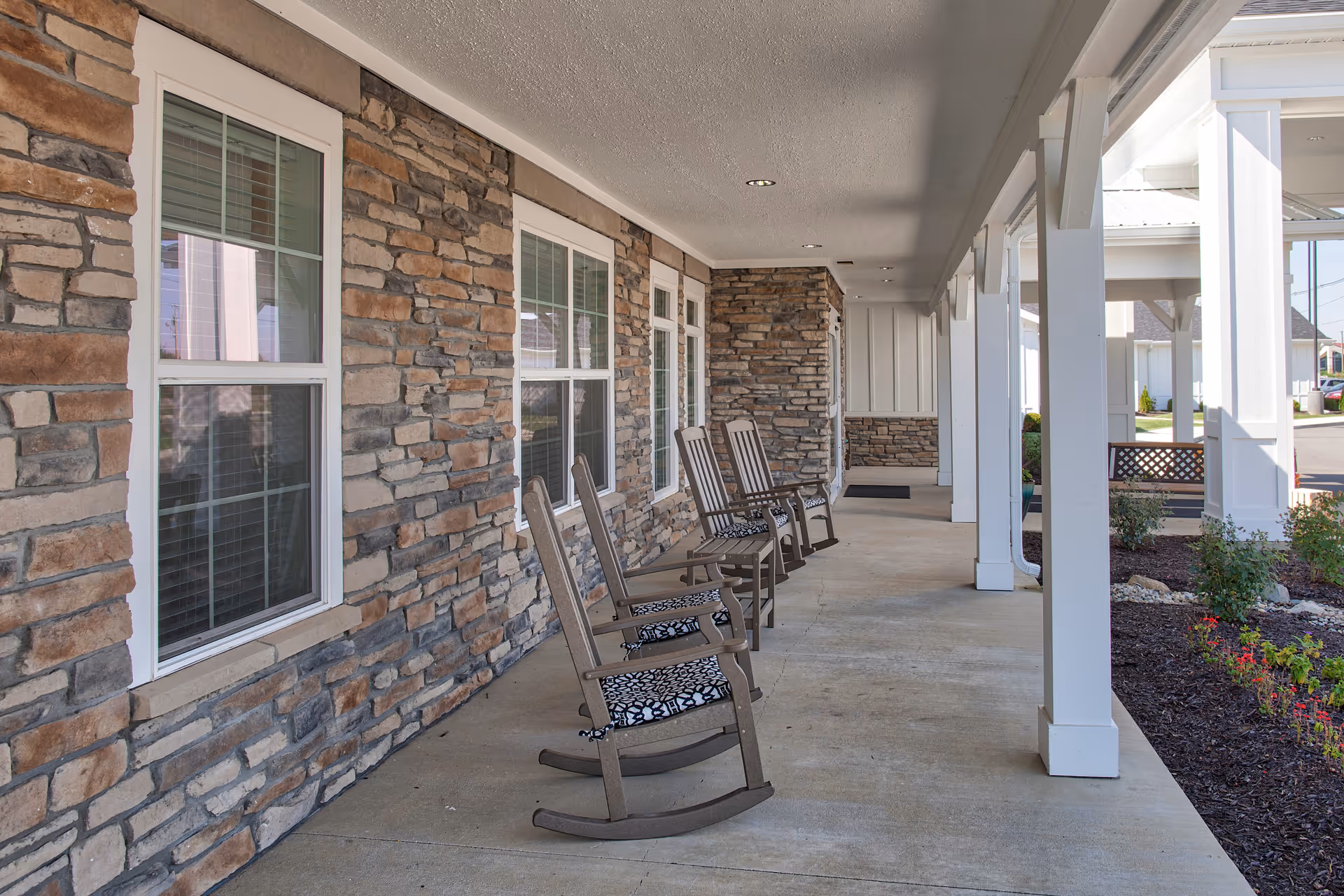 Covered outdoor porch area with stone walls and several wooden rocking chairs with patterned cushions lined up along the wall. The porch has white pillars and overlooks a landscaped garden bed with small bushes and flowers.