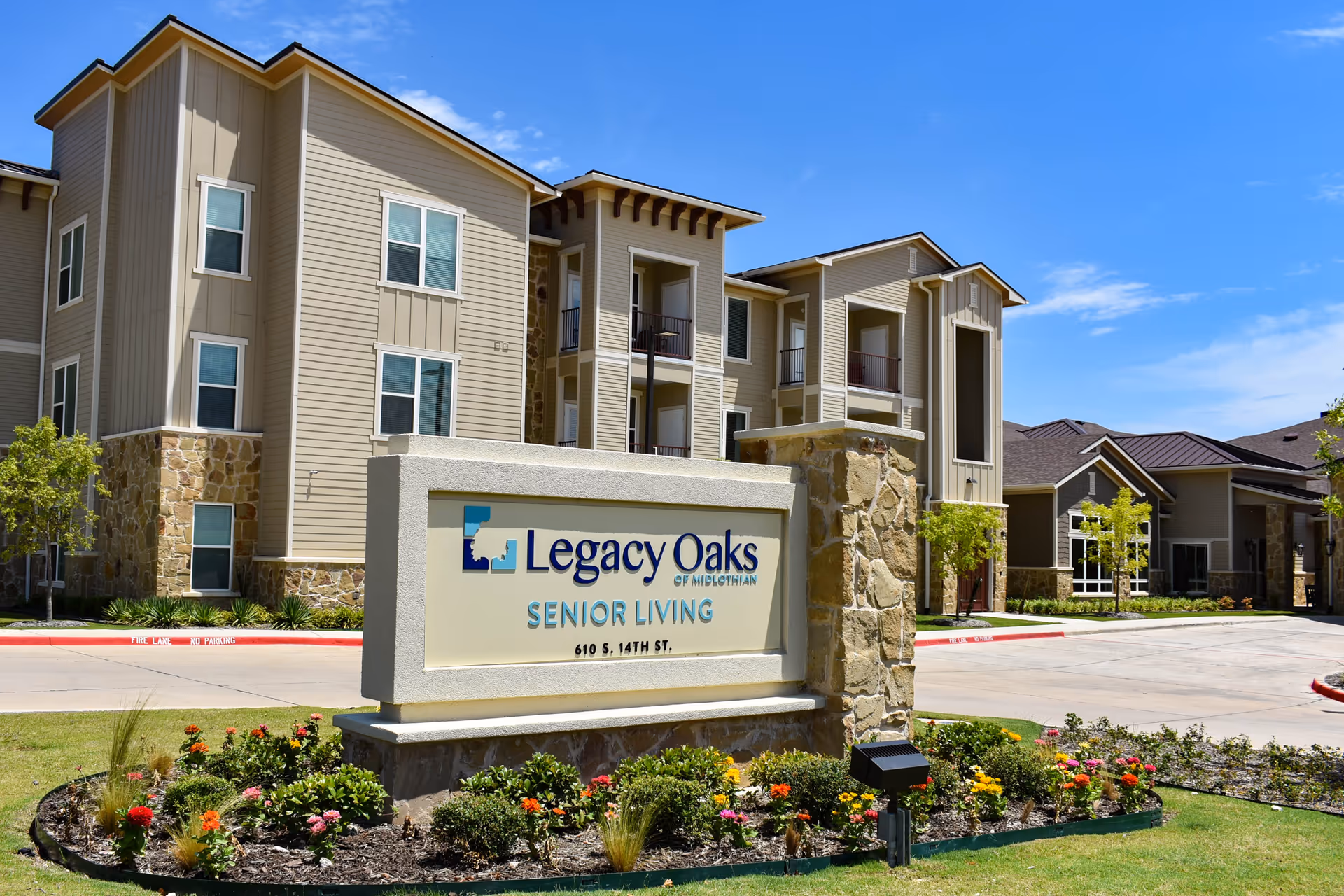 Exterior view of Legacy Oaks of Midlothian Senior Living facility showing a multi-story building with beige siding and stone accents under a clear blue sky. In the foreground is a large sign with the facility's name and address, surrounded by a landscaped garden with colorful flowers.