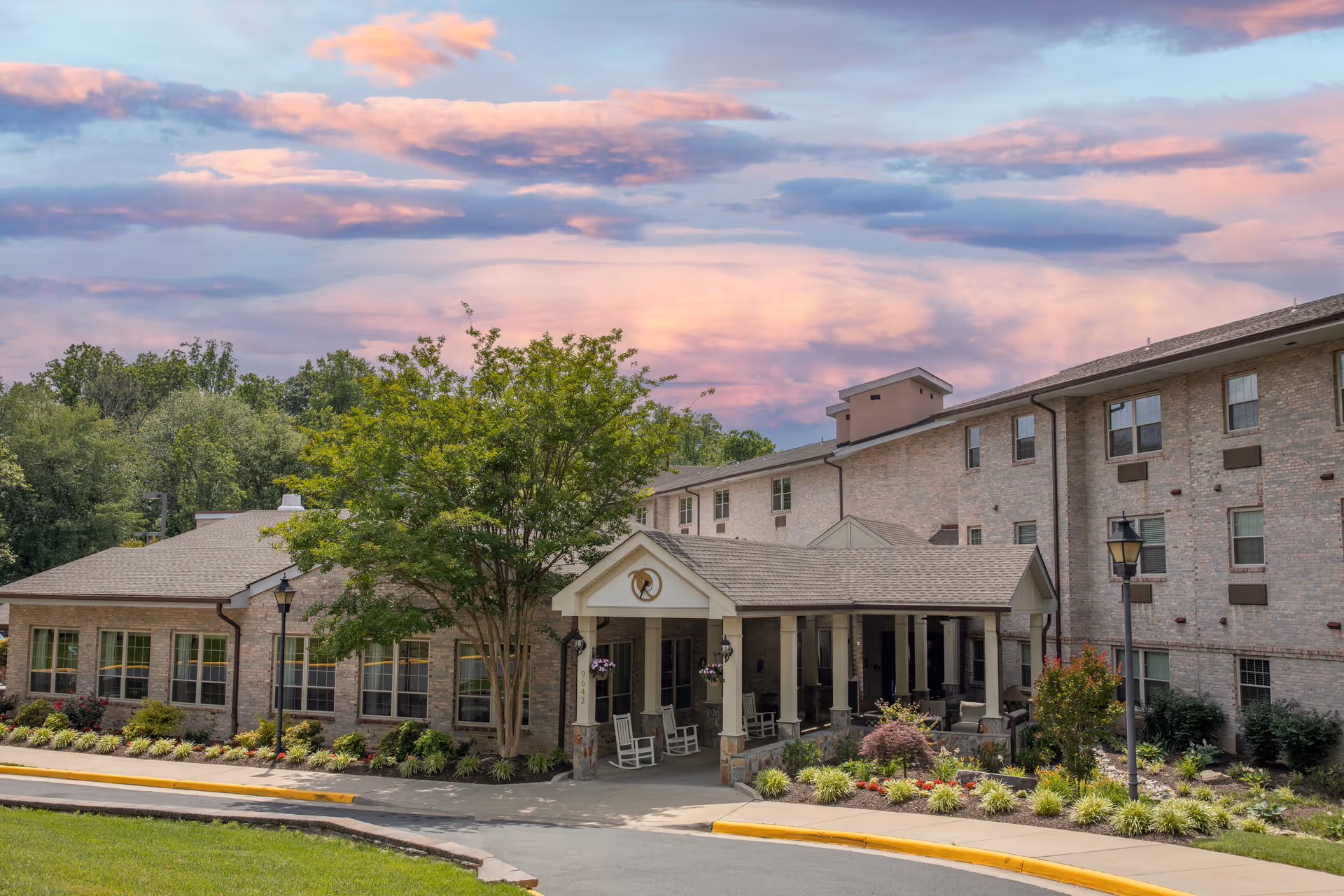 Exterior view of Heatherwood Retirement Community building at sunset with a covered entrance, rocking chairs on the porch, well-maintained landscaping, and a colorful sky with pink and purple clouds.