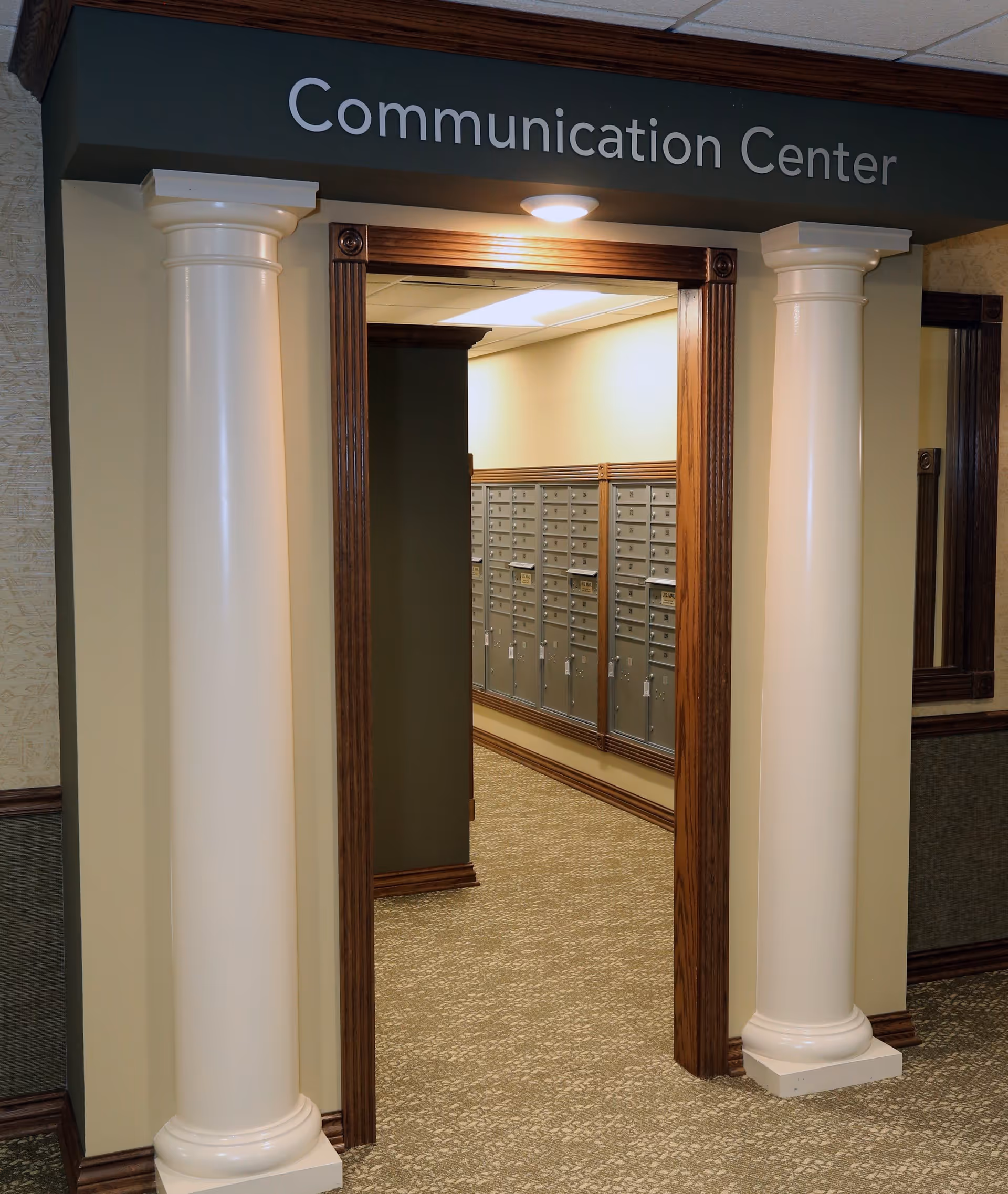 Entrance to the Communication Center in Village Crossing Retirement Community, featuring two white decorative columns on either side of a wooden door frame. Inside, there are multiple rows of mailboxes along the wall and a ceiling light above the doorway.