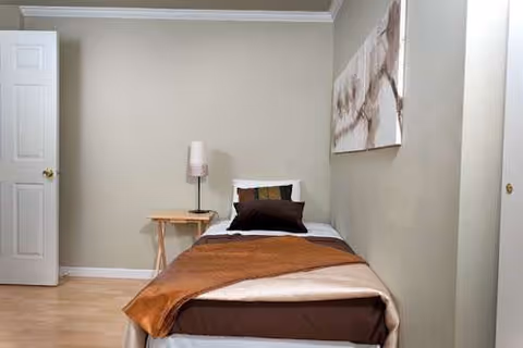 Small bedroom with single bed, brown blanket, side table lamp, and wall art.
