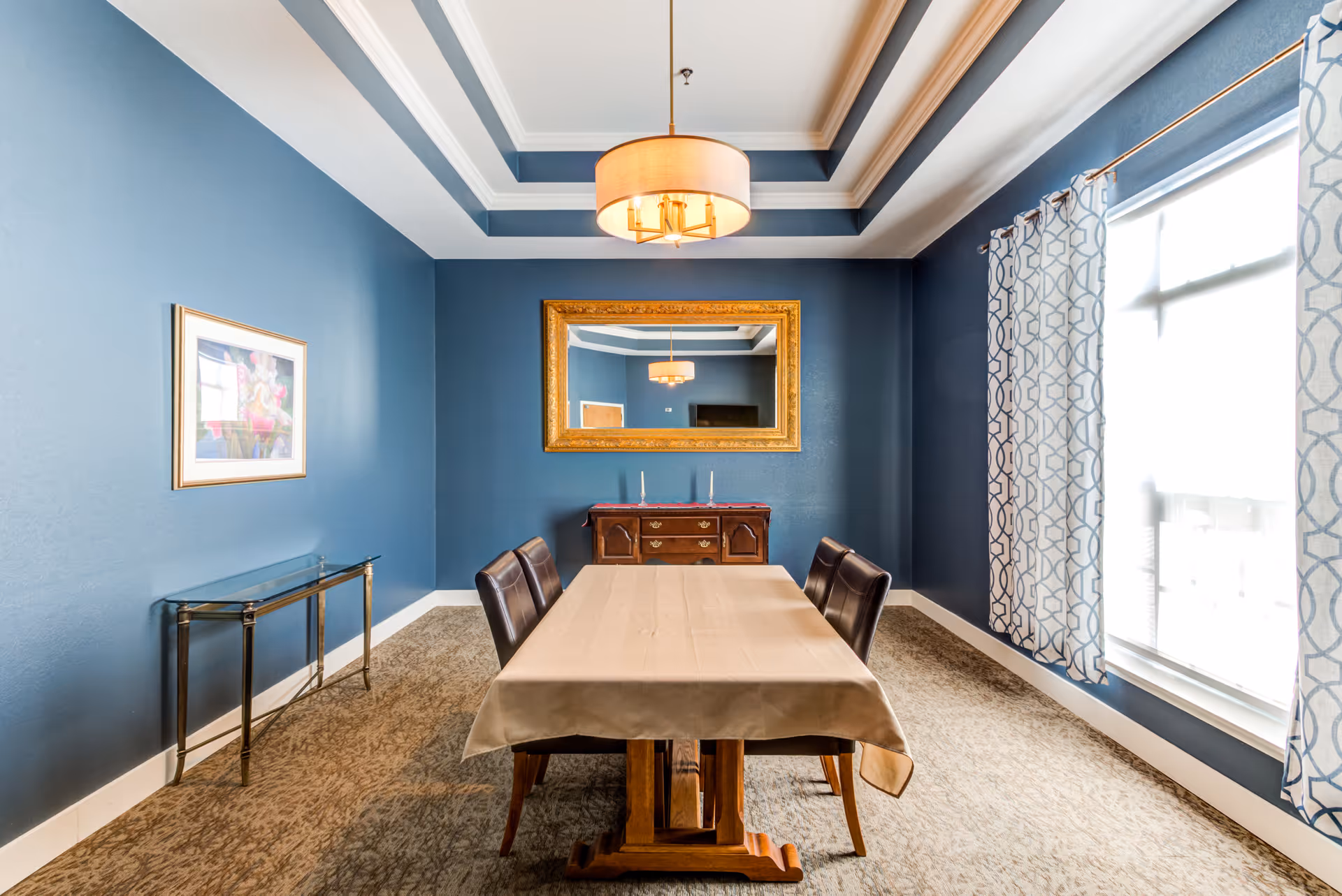 A dining room with blue walls and a beige carpet. There is a rectangular wooden dining table covered with a beige tablecloth and four dark brown leather chairs. A large gold-framed mirror hangs on the wall above a wooden sideboard with two candlesticks. A modern ceiling light fixture hangs above the table. A window with patterned curtains allows natural light into the room. A framed picture is on the left wall and a glass console table is beneath it.