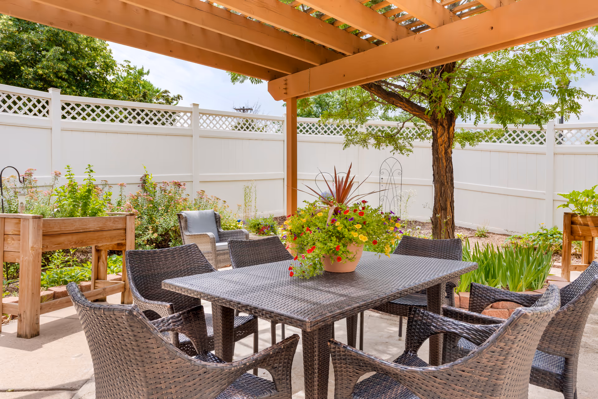 Outdoor patio area with a wicker dining table and six matching chairs under a wooden pergola. A large potted plant with colorful flowers is placed on the table. Surrounding the patio are raised garden beds, a tree, and a white fence with lattice trim. Green plants and shrubs are visible in the garden beds.