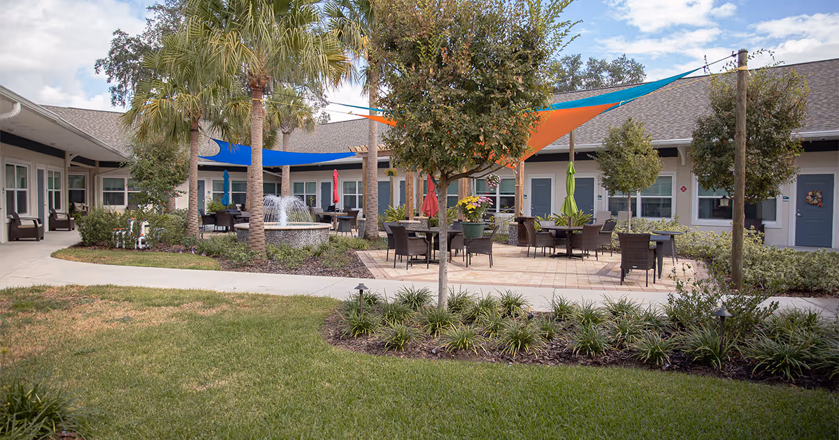 Outdoor courtyard area of an assisted living facility with palm trees, a central water fountain, tables and chairs with colorful umbrellas and shade sails, surrounded by a single-story building with multiple doors and windows.