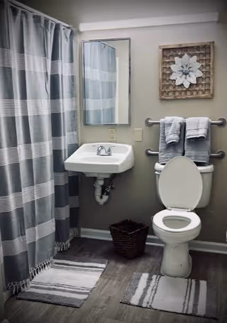 A bathroom with a white toilet with the lid open, a white wall-mounted sink with a chrome faucet, a rectangular mirror above the sink, and a gray and white striped shower curtain. Two gray towels hang on a double towel bar above the toilet. There is a decorative wall piece with a white flower above the towels. The floor has two gray and white striped rugs and a brown woven basket is placed between the sink and toilet.