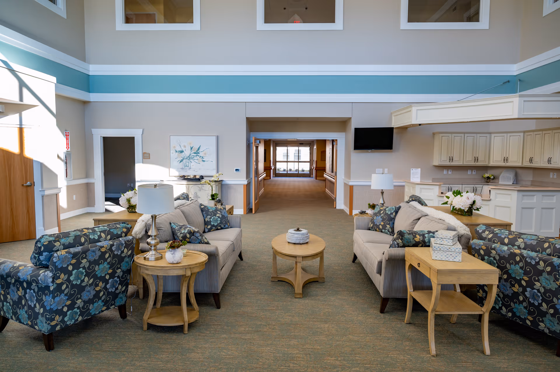 A bright and spacious common area in a senior living facility featuring two beige sofas and two floral armchairs arranged around two wooden coffee tables. The room has a high ceiling with windows near the top, light-colored walls with a blue accent stripe, and a hallway leading to another part of the building. There is a flat-screen TV mounted on the wall above a white counter with cabinets, and decorative flowers and lamps on the tables.