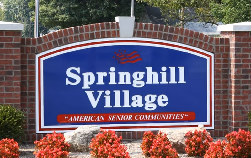 A large blue and white sign with red accents reading 'Springhill Village' and 'American Senior Communities' mounted on a brick wall with red flowers and rocks in front, surrounded by trees and greenery.