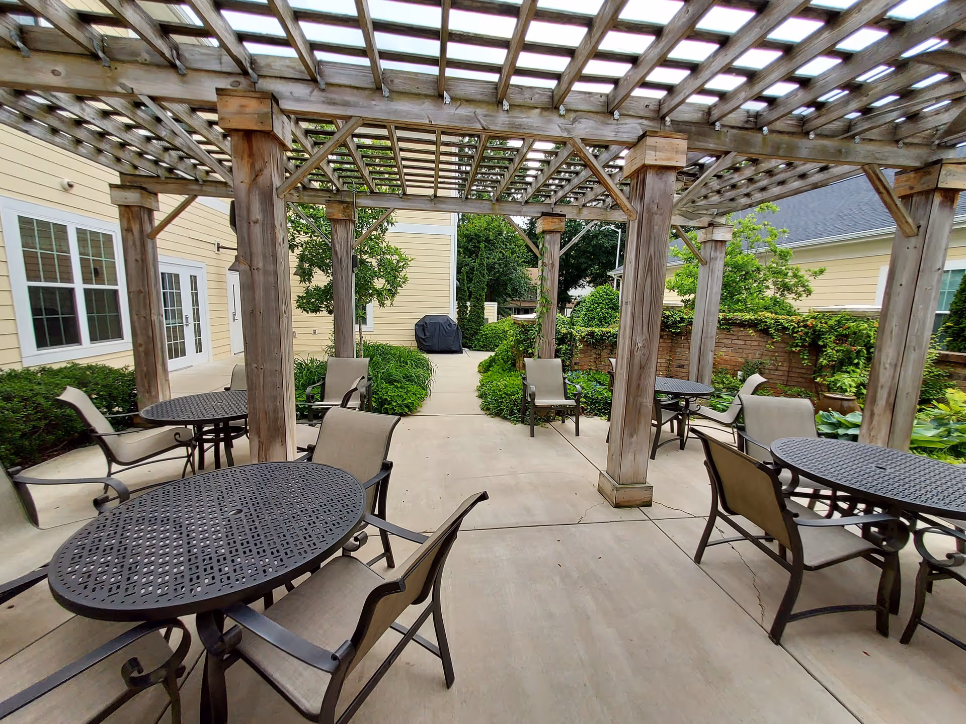 Outdoor patio area with several round metal tables and beige cushioned chairs under a wooden pergola. The patio is surrounded by greenery, bushes, and a yellow building with windows and doors in the background.
