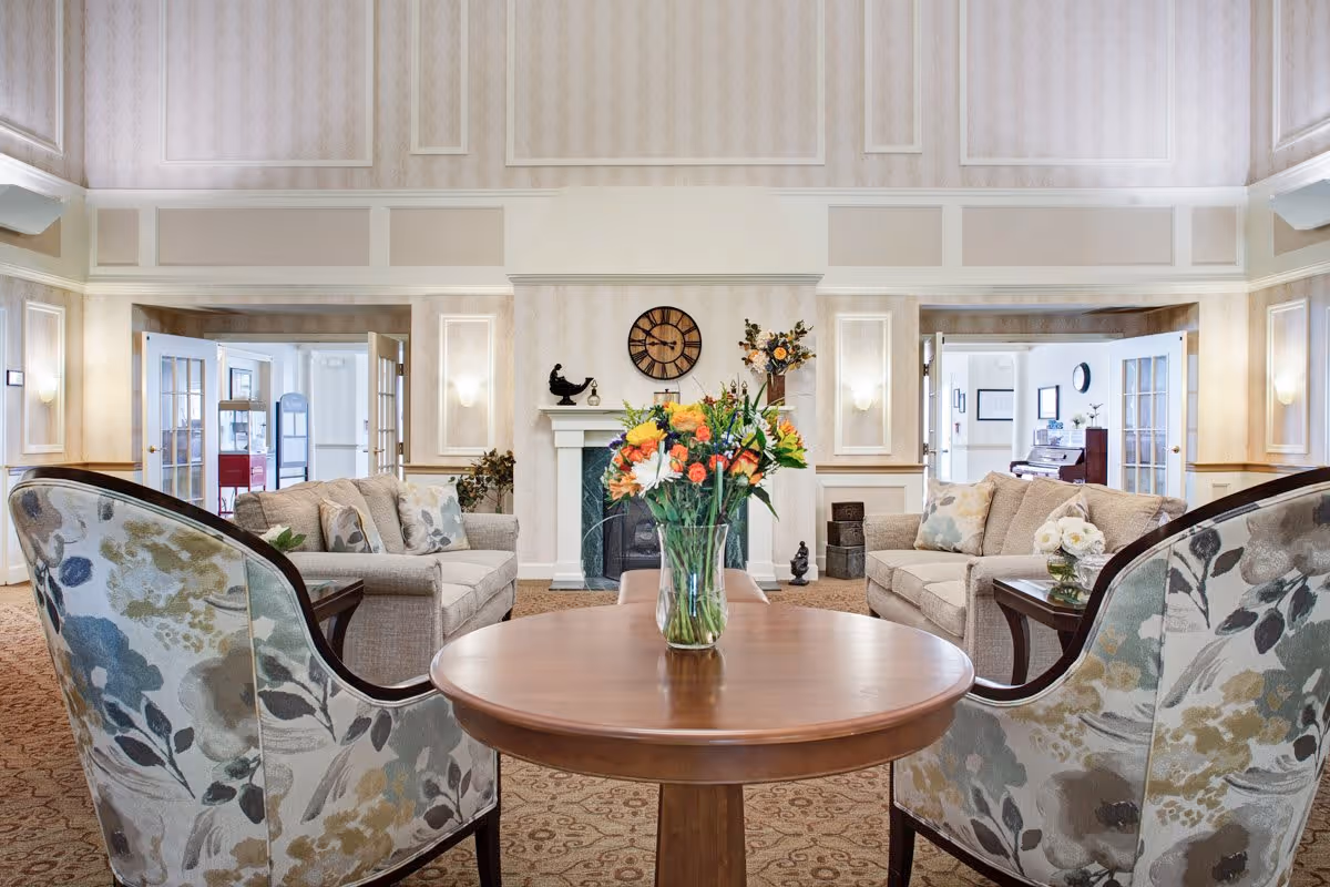 A bright and elegant living room area in a senior living facility featuring two floral-patterned armchairs and two beige sofas arranged around a round wooden table with a vase of colorful flowers. The room has a fireplace with a clock and decorative items above it, soft wall lighting, and double glass doors leading to other rooms.