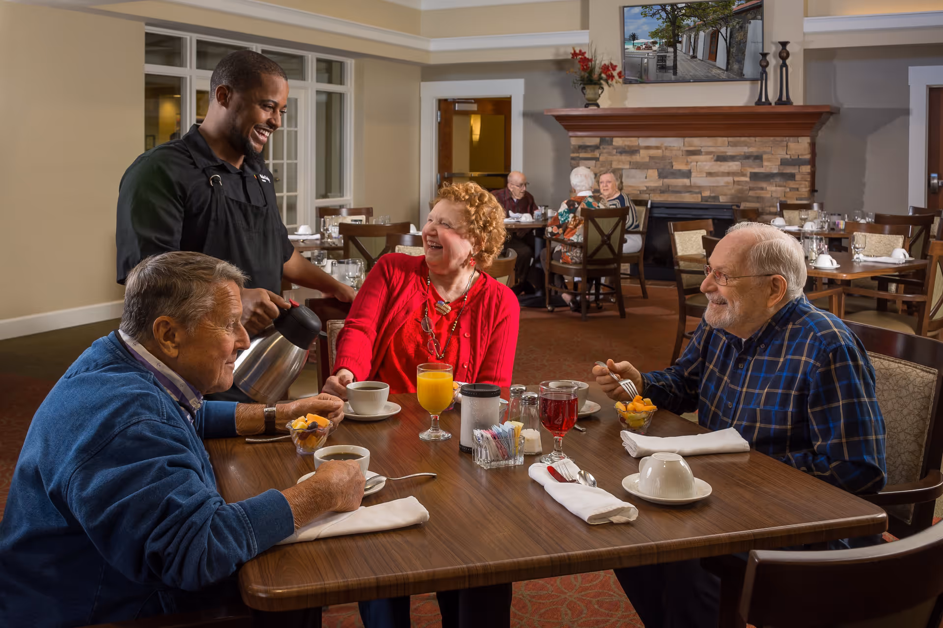A smiling waiter pours coffee for three elderly people seated around a dining table in a cozy dining room. The table is set with cups, glasses of juice, bowls of fruit, and napkins. In the background, other elderly people are seated at tables near a stone fireplace with a painting above it.