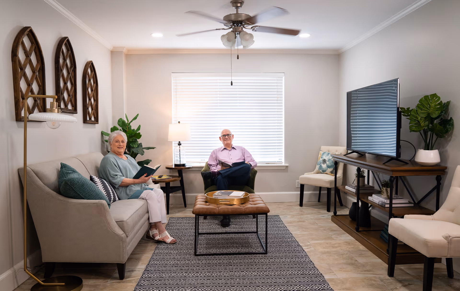 A cozy living room in a senior living facility with two elderly people sitting and reading. The room features a beige sofa with green and patterned pillows, a green armchair, a brown tufted ottoman, a TV on a wooden stand, two beige chairs, a floor lamp, a table lamp, decorative wall art, and plants. The room has a window with blinds and a ceiling fan with lights.