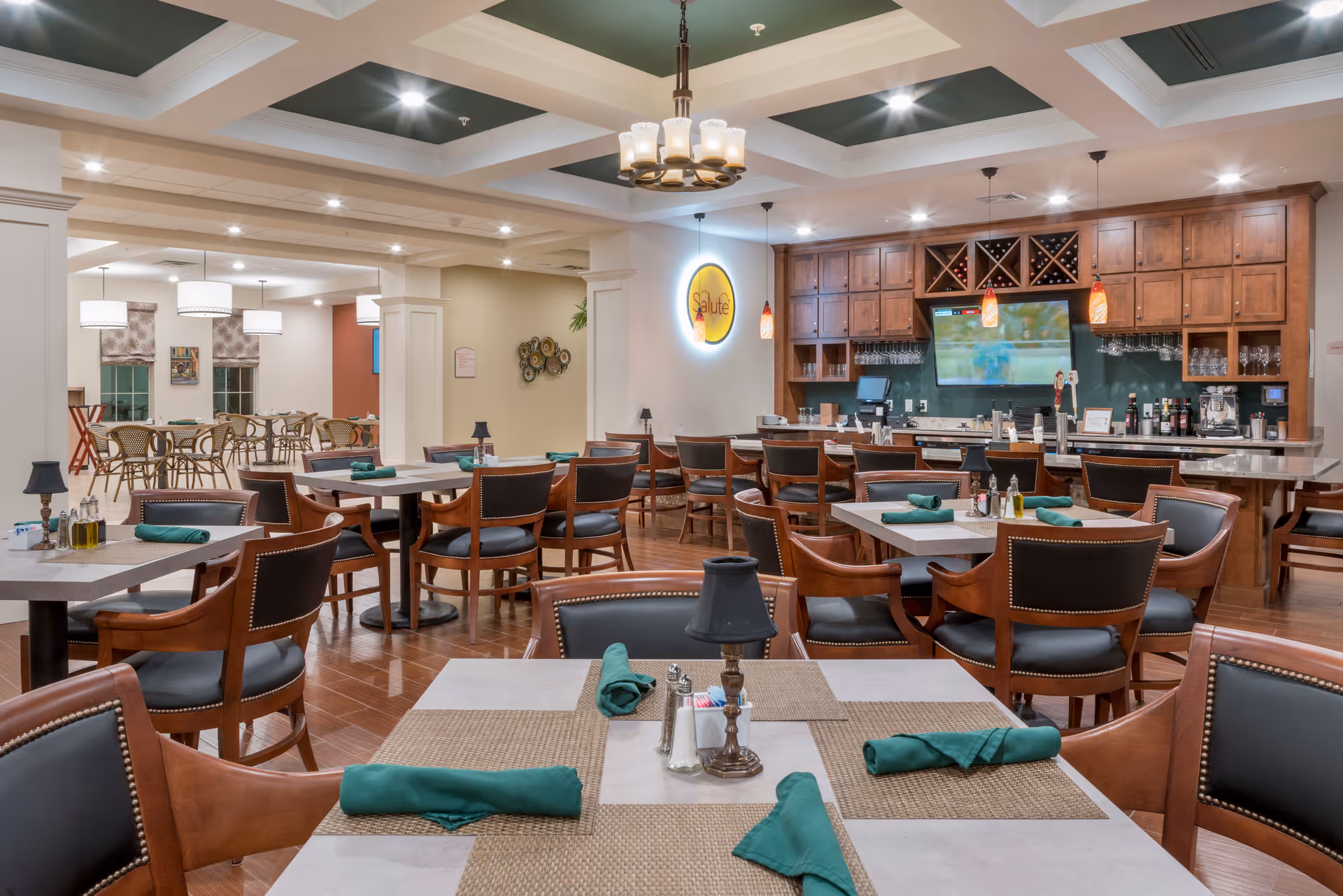 A spacious dining room with multiple tables and chairs, each table set with green napkins and placemats. The room features a bar area with wooden cabinets, wine racks, and a television mounted on the wall. The ceiling has recessed lighting and a decorative chandelier. In the background, there is another seating area with additional tables and chairs.