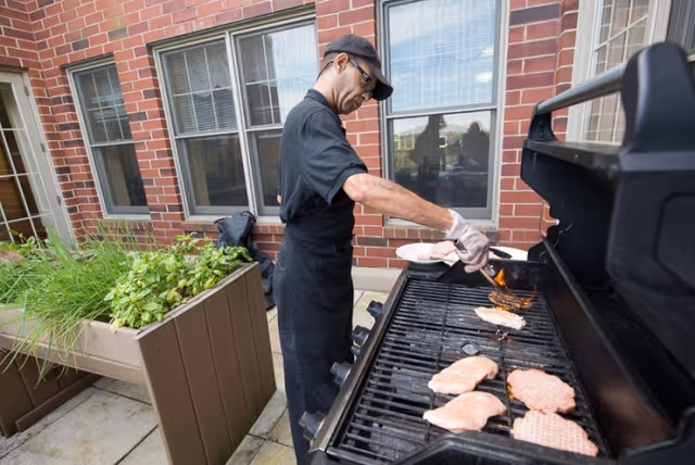 A person wearing an apron and cap grills chicken and burger patties on a gas barbecue on a courtyard patio beside planter boxes and a brick building.