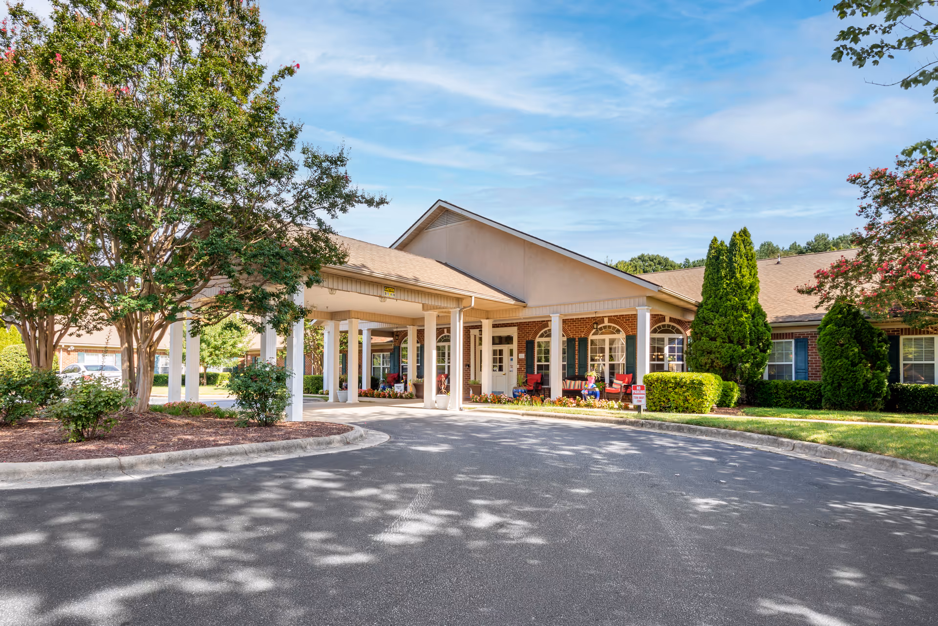 Exterior view of Brookdale Concord Parkway senior living facility showing a covered entrance with white pillars, brick walls, large windows with green shutters, and a landscaped area with trees and bushes under a blue sky.