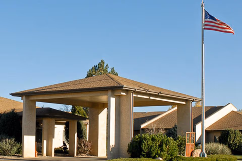 Covered entrance/porte-cochère of a senior living facility with an American flag on a flagpole and nearby shrubs.