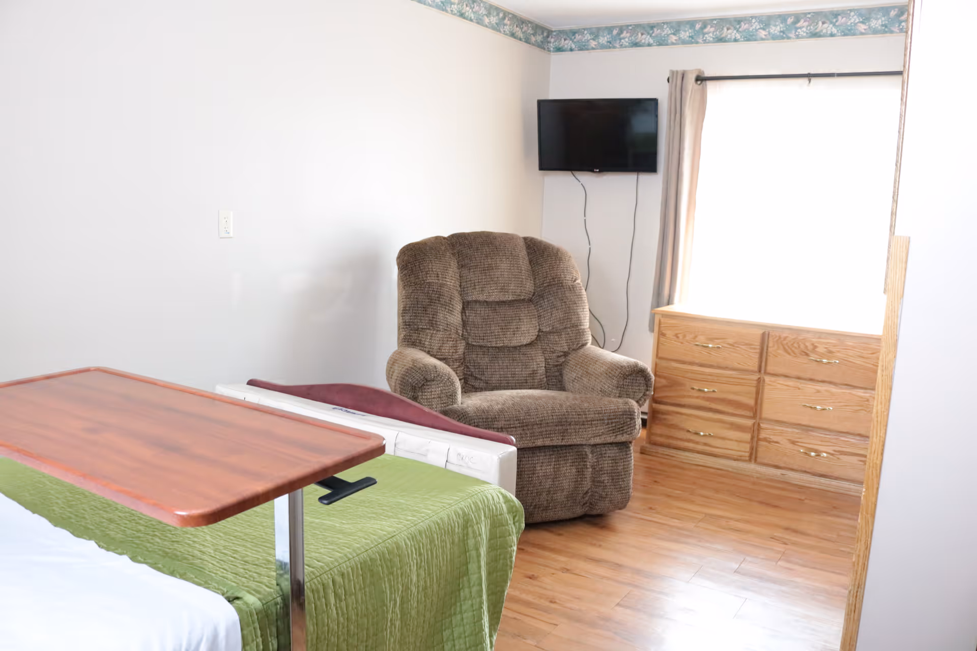 A small room with a hospital-style bed covered with a green blanket and a wooden overbed table. There is a brown cushioned recliner chair next to a window with beige curtains. A flat-screen TV is mounted on the wall above a wooden dresser with six drawers. The floor is wooden, and the walls are white with a floral wallpaper border near the ceiling.