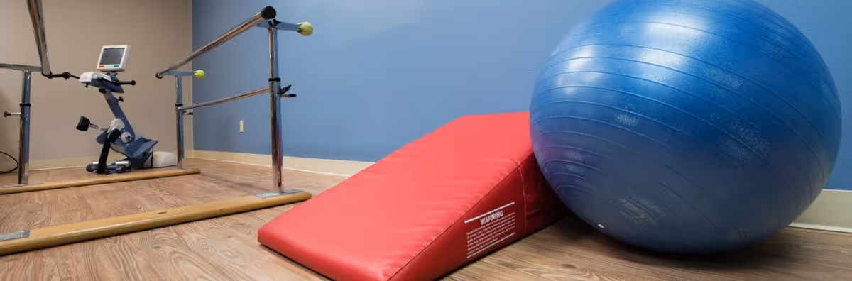 Physical therapy room with parallel bars, an exercise bike, a red wedge mat and a large blue exercise ball on a wood floor.