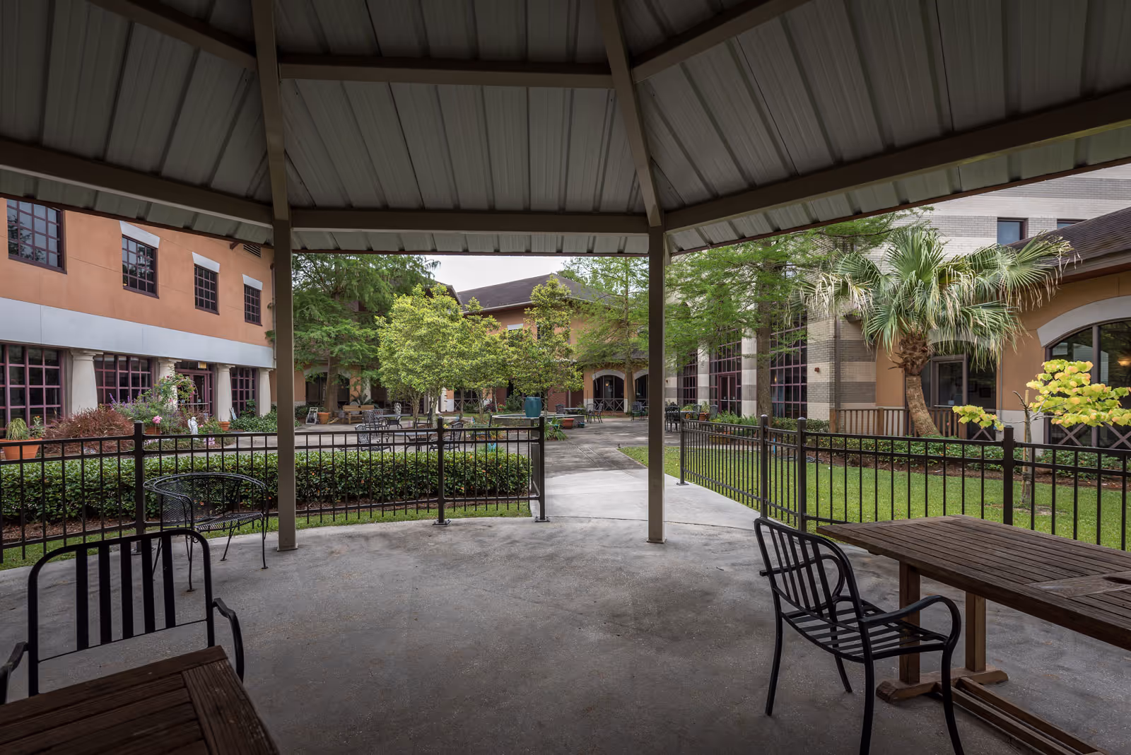 View from inside a covered outdoor pavilion looking out onto a courtyard with pathways, green grass, trees, and surrounding buildings with large windows. There are metal chairs and wooden tables under the pavilion and in the courtyard.