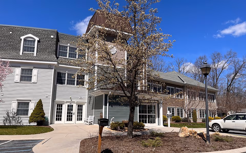 Exterior view of a senior living facility building with white and brown siding, multiple windows, and a tree with blooming flowers in front. There is a sidewalk leading to the entrance, a parking area with a white vehicle, and a lamppost near the entrance under a clear blue sky.