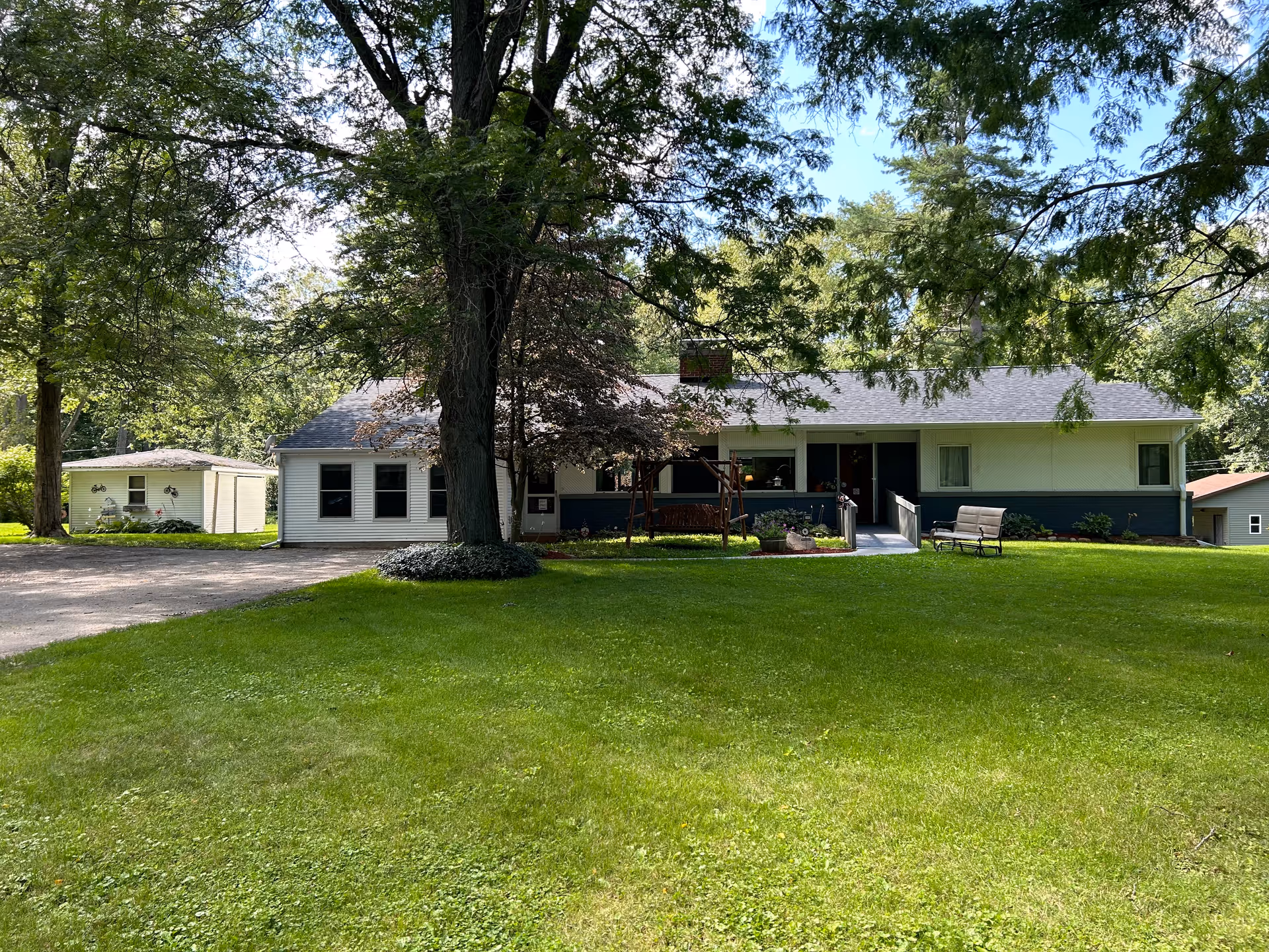 Single-story house with a front porch, swing and benches set behind a large grassy yard and shaded by trees.