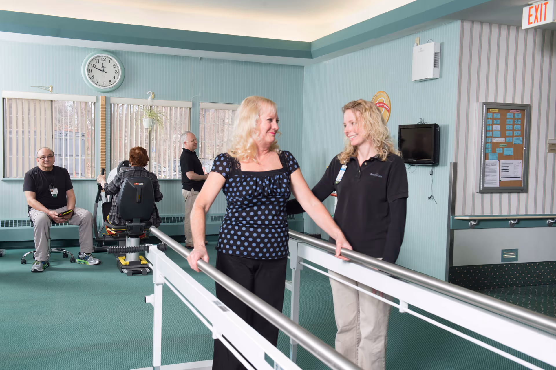A senior woman is walking with support bars assisted by a caregiver in a rehabilitation or therapy room. In the background, two other seniors are using exercise equipment, and another man is seated watching. The room has light blue walls, large windows with vertical blinds, a clock, and an exit sign.