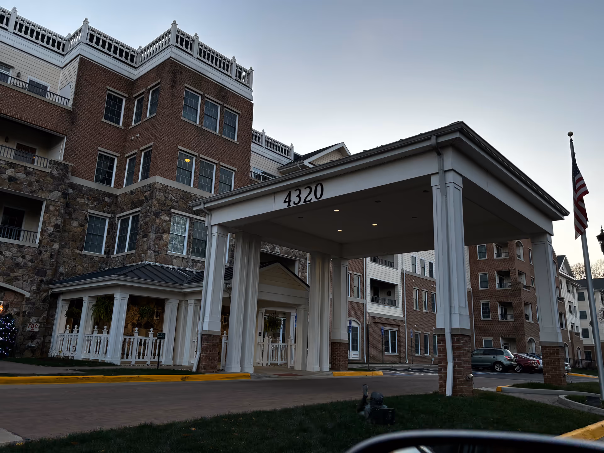 Exterior view of a multi-story senior living facility building with a covered entrance displaying the number 4320. The building features brick and stone facades with multiple windows and balconies. An American flag is visible on the right side near the entrance driveway.