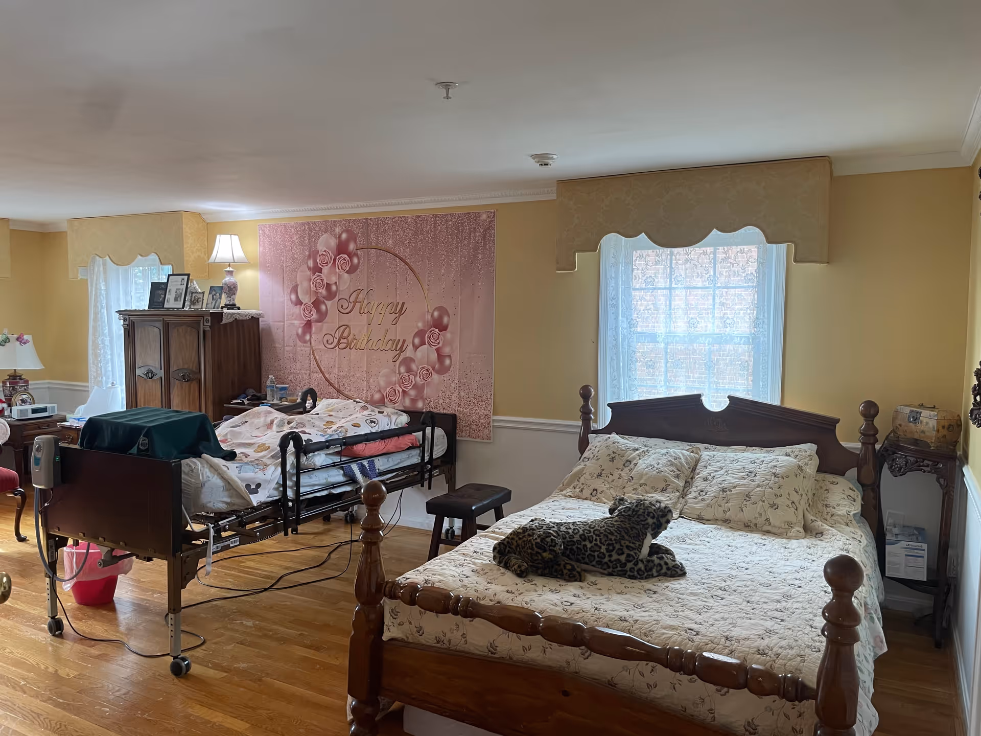 A bedroom in an assisted living facility with two beds. One bed is a hospital-style bed with a person lying under a blanket, and the other is a wooden bed with floral bedding and a stuffed leopard toy on it. The room has wooden floors, two windows with sheer curtains and valances, a wooden cabinet with framed photos and a lamp, and a 'Happy Birthday' banner on the wall.