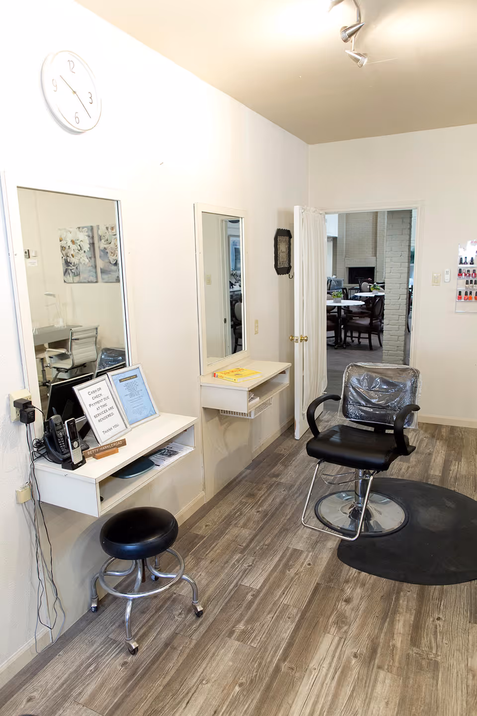 Interior view of a small salon area with a black salon chair on a black mat, a black rolling stool, two wall-mounted mirrors with small shelves underneath, a clock on the wall, and a door leading to another room with a fireplace and dining table visible.