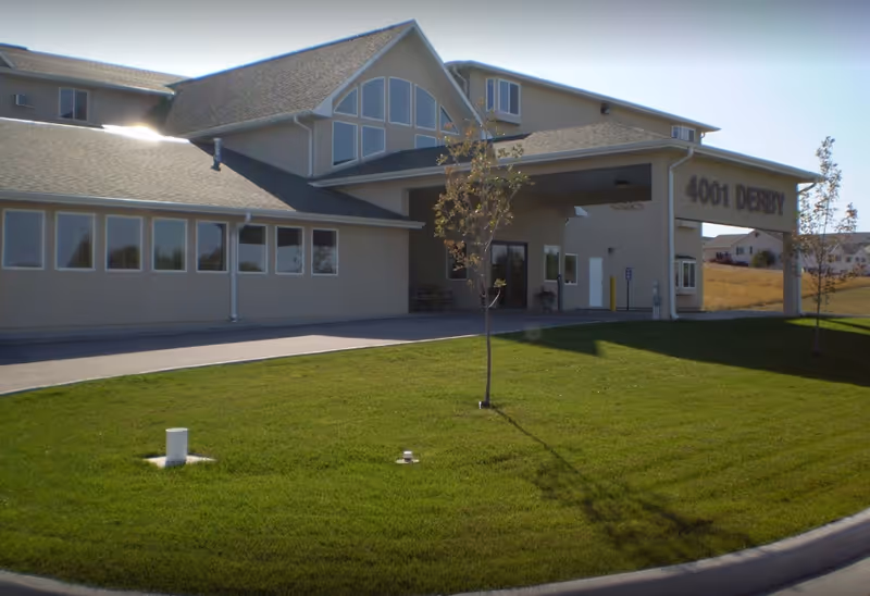 Exterior view of a single-story building with a covered entrance and large windows. The building has a beige facade and a sloped roof. There is a well-maintained green lawn with a small tree in front. The address '4001 DERBY' is visible on the building near the entrance.