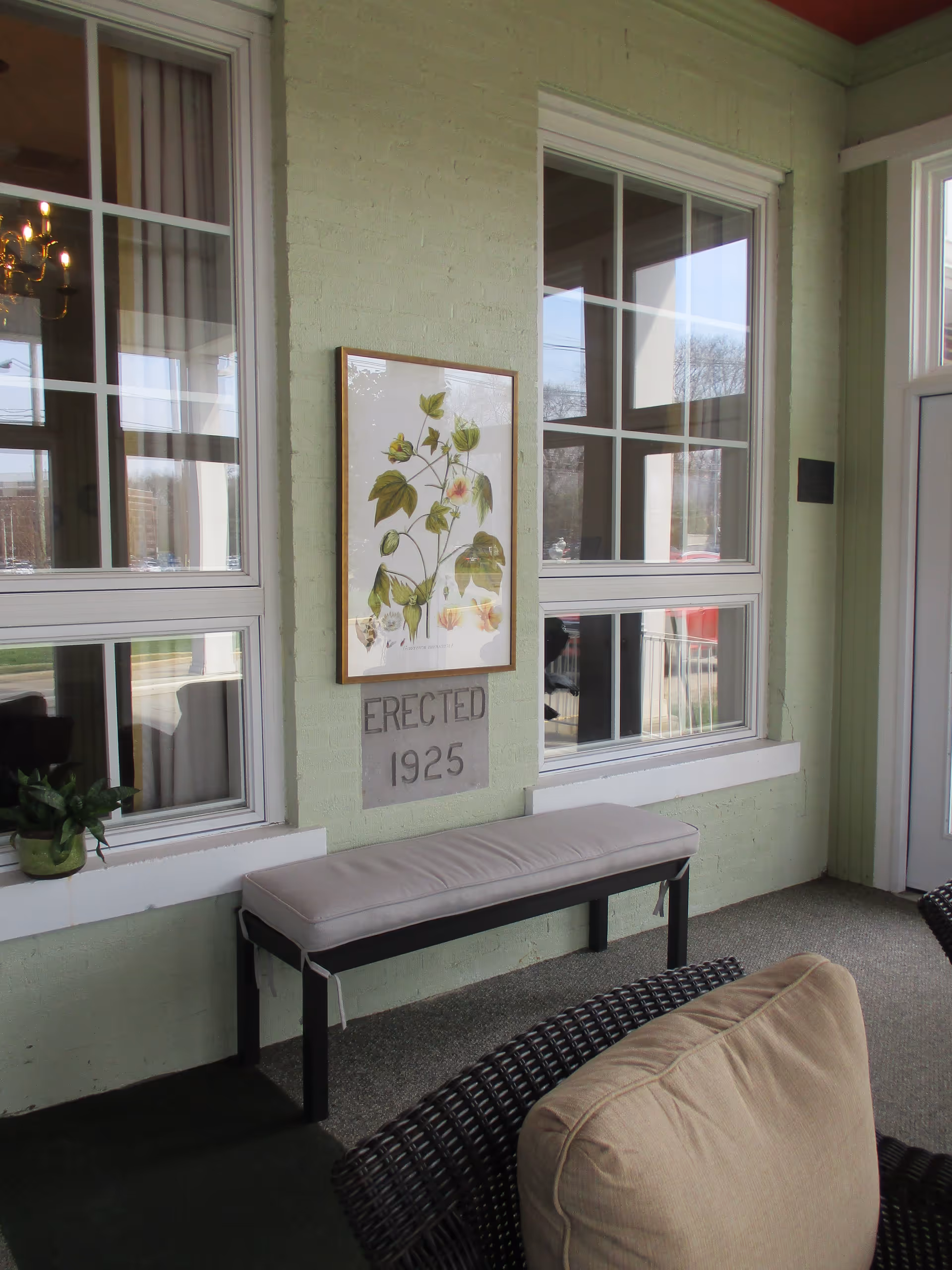 Indoor seating area with a cushioned bench and a wicker chair with a beige cushion. The walls are light green with two large windows and a framed botanical print hanging above a stone plaque that reads 'ERECTED 1925'. A small potted plant sits on the windowsill.