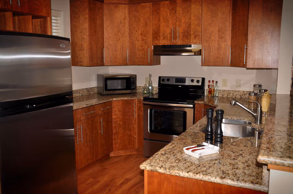 A modern kitchen with wooden cabinets, granite countertops, a stainless steel refrigerator, an electric stove with oven, a microwave, a sink with a faucet, and various kitchen items including salt and pepper shakers and condiment bottles.
