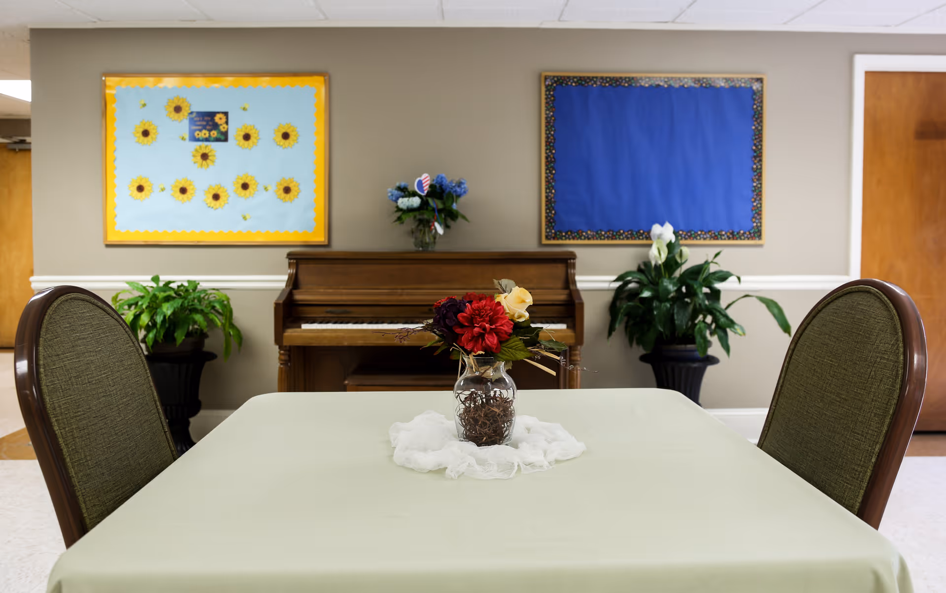 A small table covered with a light green tablecloth with a vase of artificial flowers in the center, two green cushioned chairs on either side, a wooden piano against the wall behind the table, two potted plants on either side of the piano, and two bulletin boards on the wall, one decorated with sunflowers and the other blank with a blue background.